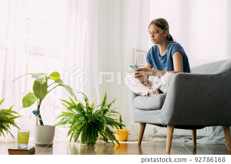 A young girl is sitting in a chair and exchanging messages online on her phone. Home recreation, modern lifestyle A young girl is sitting in a chair and exchanging messages online on her phone. Home recreation, modern lifestyle 92786168