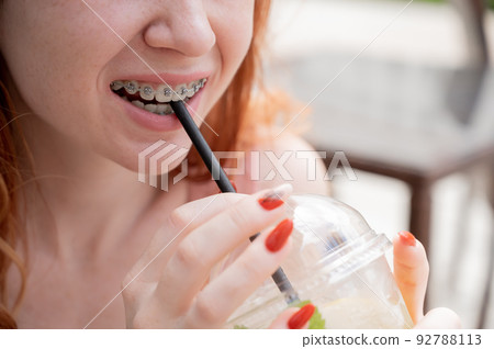 Young beautiful red-haired woman with braces drinks cooling lemonade outdoors in summer. Portrait of a smiling girl with freckles. Young beautiful red-haired woman with braces drinks cooling lemonade outdoors in summer. Portrait of a smiling girl with freckles. 92788113