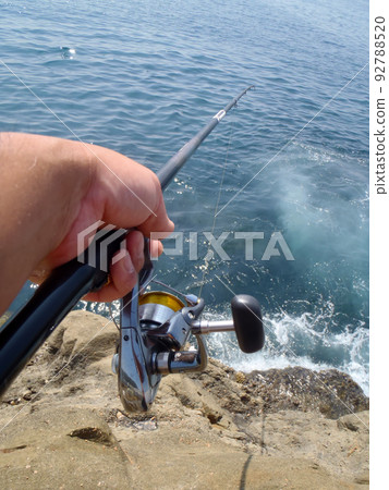A fisherman's hand sticking out a high-quality rod from the surf fishing spot to the sea and the scenery of the salted fish that stretches out to sea. Taken under sunny summer weather. 92788520