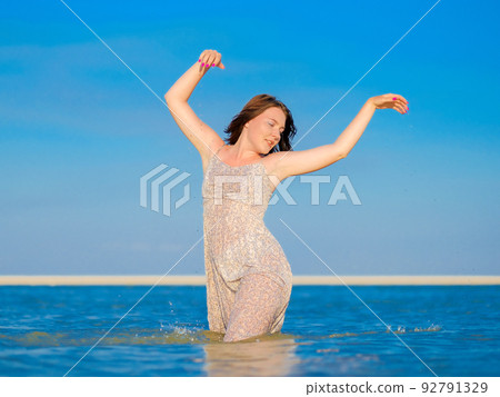 A young woman in a wet sundress enjoys splashing water while standing knee-deep in the sea 92791329