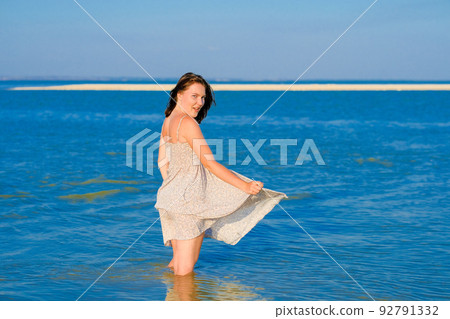 A young girl in a sundress fluttering in the wind stands knee-deep in the sea 92791332