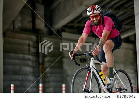 Portrait of a cyclist standing under a bridge with a bicycle in his hand, posing at the camera against an architecture background. Active lifestyle. Cycling is a hobby. 92796642