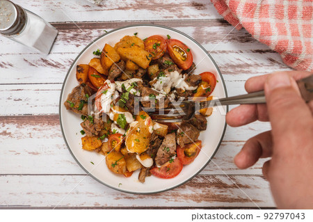 Hand taking a portion of beef chunks sauteed with cassava or manioc, chives, pepper, lemon and cherry tomato on a rustic surface. 92797043