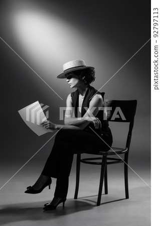 Portrait of stylish woman in classic black suit and straw hat, posing, sitting on chair and reading. Black and white photography Portrait of stylish woman in classic black suit and straw hat, posing, sitting on chair and reading. Black and white photography 92797113