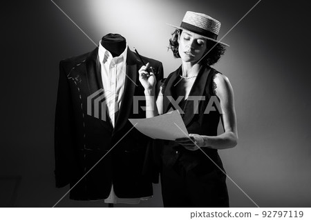 Portrait of talented woman, fashion designer standing near dummy with stylish classical male suit. Black and white photography. Working with pattern 92797119