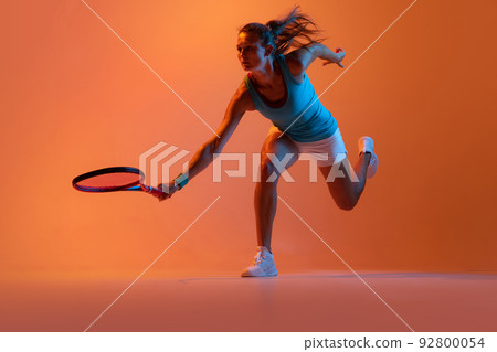 Dynamic portrait of woman, professional tennis player training isolated over orange studio background in neon light Dynamic portrait of woman, professional tennis player training isolated over orange studio background in neon light 92800054