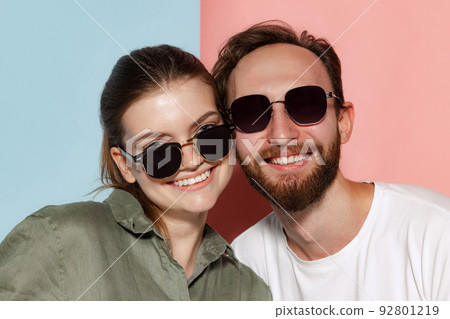 Closeup young bearded man and beautiful girl in sunglasses looking at camera posing isolated over blue-pink background. Emotions, youth, facial expressions Closeup young bearded man and beautiful girl in sunglasses looking at camera posing isolated over blue-pink background. Emotions, youth, facial expressions 92801219
