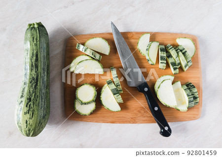 Cutting zucchini on wooden board, whole squash, knife and slices, overhead view 92801459