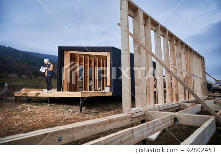Male architect building wooden frame house in the Scandinavian style barnhouse. Man builder standing on construction site in safety helmet, with construction documentation inspecting quality of work. 92802451