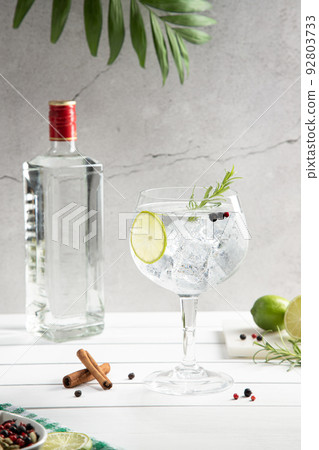 Cold glass of gin tonic and bottle on white wooden base on light background. Vertical format. Cold glass of gin tonic and bottle on white wooden base on light background. Vertical format. 92803733
