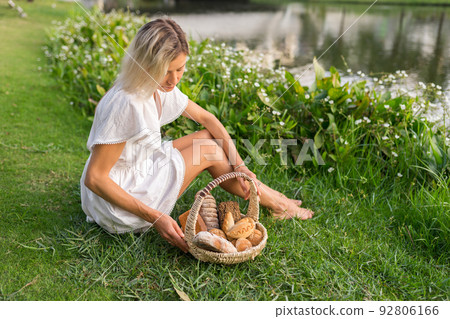 Young woman in white dress sitting on the grass with basket with assorti of brown and white homemade bread. High quality photo 92806166