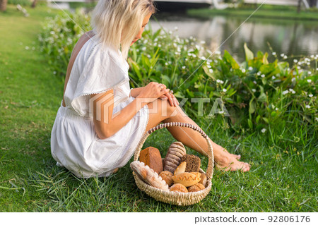 Young woman in white dress sitting on the grass with basket with assorti of brown and white homemade bread. High quality photo 92806176