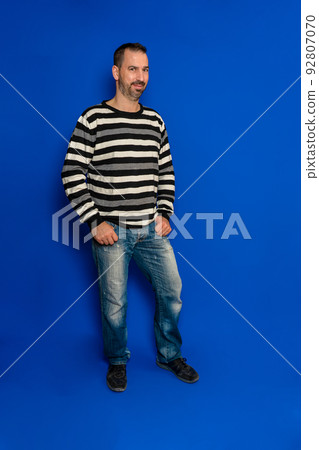 Tall spanish man with a beard wearing a striped sweater and jeans standing isolated over blue background with his hands in his pockets. Tall spanish man with a beard wearing a striped sweater and jeans standing isolated over blue background with his hands in his pockets. 92807070