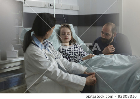 Pediatrician examining sick girl disease symptoms while resting in patient bed. Doctor expert discussing with girl about her health condition while caring father sitting beside ill daughter. Pediatrician examining sick girl disease symptoms while resting in patient bed. Doctor expert discussing with girl about her health condition while caring father sitting beside ill daughter. 92808816