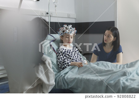 Neurology specialist examining neurological disease state of ill kid. Hospitalized sick girl wearing EEG headset resting in patient bed while doctor analyzing brain condition. 92808822