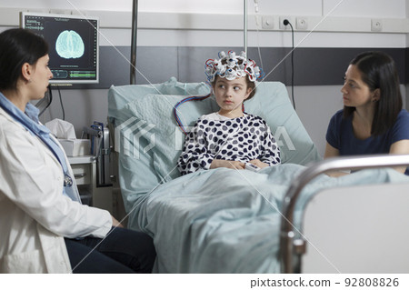 Neurology specialist measuring neurologic disease evolution of sick kid sitting in pediatric healthcare facility. Expert pediatrician monitoring hospitalized girl brain condition using EEG headset. Neurology specialist measuring neurologic disease evolution of sick kid sitting in pediatric healthcare facility. Expert pediatrician monitoring hospitalized girl brain condition using EEG headset. 92808826