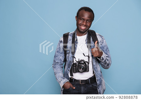Young photographer showing thumbs up hand gesture while having professional camera on blue background. Happy photography enthusiast with DSLR photo device giving okay approve hand symbol. 92808878