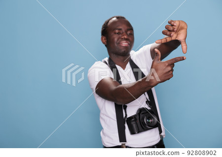 Young guy with DSLR device gesturing taking photo with hands. Confident professional photographer doing frame gesture with hands while having modern camera on blue background. Studio shot 92808902