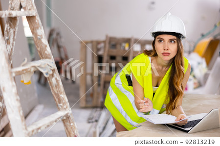 Female foreman in a protective helmet and a yellow vest checks the execution of repair work using laptop Female foreman in a protective helmet and a yellow vest checks the execution of repair work using laptop 92813219