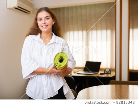 Cheerful young female entrepreneur standing with yoga matt in office 92813348