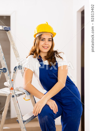 Portrait of positive builder woman in blue overalls next to stepladder 92813402