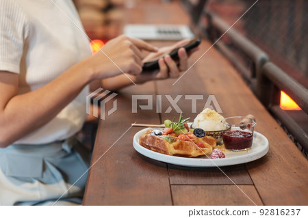 Woman using smartphone and eating dessert with fresh berries and fruit at cafe 92816237