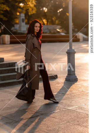 Outdoor fashion portrait of young elegant fashionable brunette woman, model in stylish hat, choker and light raincoat posing at sunset in European city. Outdoor fashion portrait of young elegant fashionable brunette woman, model in stylish hat, choker and light raincoat posing at sunset in European city. 92816389
