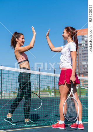 Two Caucasian women in sportswear greeting before a tennis match on an outdoor court. Players give a high five before the game. 92820055