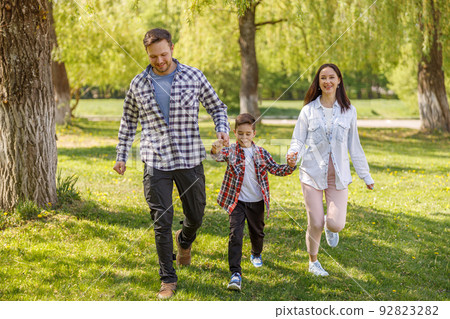 Cheerful parents with small son walking hand in hand in green summer park. Happy family. Cheerful parents with small son walking hand in hand in green summer park. Happy family. 92823282