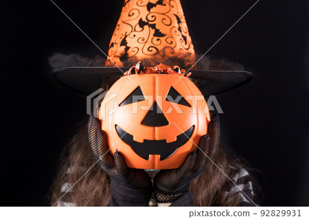 A woman in a witch costume in a hat holds a bucket of candy instead of a head. Halloween people. A woman in a witch costume in a hat holds a bucket of candy instead of a head. Halloween people. 92829931