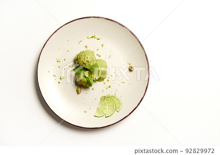 Flat lay. Food still life of three frozen green balls of pistachio and lime ice cream sorbet,sprinkled with pistachio nuts, melting on a plate, isolated on white background with copy space for text. Flat lay. Food still life of three frozen green balls of pistachio and lime ice cream sorbet,sprinkled with pistachio nuts, melting on a plate, isolated on white background with copy space for text. 92829972