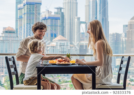 Happy family having breakfast on the balcony. Breakfast table with coffee fruit and bread croisant on a balcony against the backdrop of the big city Happy family having breakfast on the balcony. Breakfast table with coffee fruit and bread croisant on a balcony against the backdrop of the big city 92833376