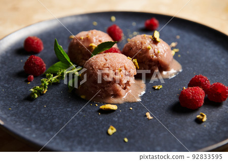Cropped view of frozen balls with homemade, dairy free berry sorbet, garnished with grated pistachios, raspberries and basil lemon leaves on blue navy plate. Food still life. Selective focus. Close-up 92833595