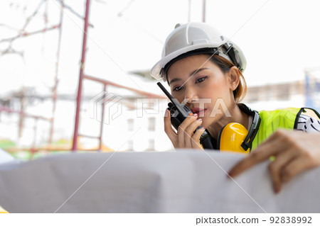 Asian engineer or Young woman Architect put on a helmet for safety and look at Blueprint for Inspect Building factory Construction Site and use walkie talkie for a talk with a contractor. Asian engineer or Young woman Architect put on a helmet for safety and look at Blueprint for Inspect Building factory Construction Site and use walkie talkie for a talk with a contractor. 92838992