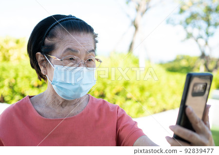 Happy Asian elderly wearing a face mask and answering a Video Call Using her Smartphone in the park and chatting with family, Technology and older people concept. Happy Asian elderly wearing a face mask and answering a Video Call Using her Smartphone in the park and chatting with family, Technology and older people concept. 92839477