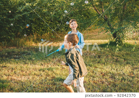 Boys play with soap bubbles in a summer park, Among the greenery. Happy children catch soap bubbles. Boys play with soap bubbles in a summer park, Among the greenery. Happy children catch soap bubbles. 92842370