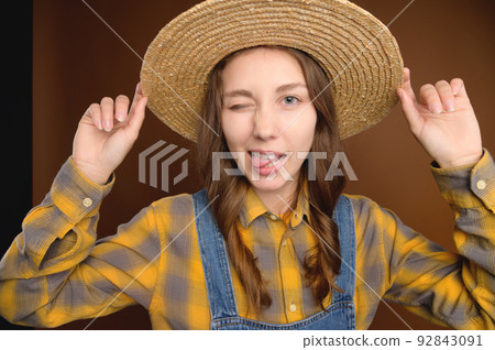 Young cute smiling female student in a straw hat and a plaid shirt, shows her tongue and winks. studio portrait Young cute smiling female student in a straw hat and a plaid shirt, shows her tongue and winks. studio portrait 92843091