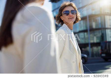 Two happy woman in trendy outfits walk outside on background of modern building Two happy woman in trendy outfits walk outside on background of modern building 92843349