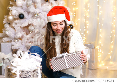 Happy caucasian woman in santa claus hat and light sweater holds a gift box 92843984