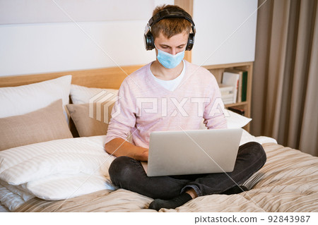 young man in headphones and protective mask at home with laptop 92843987