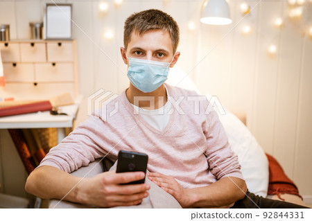 a young man sits on a sofa wearing a protective mask with a phone in his hand a young man sits on a sofa wearing a protective mask with a phone in his hand 92844331