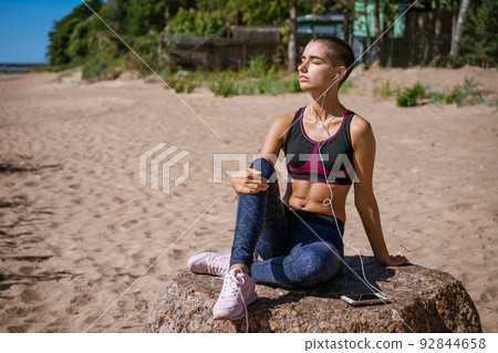 Young woman dressed in sportswear sits on beach for learning music listening Young woman dressed in sportswear sits on beach for learning music listening 92844658