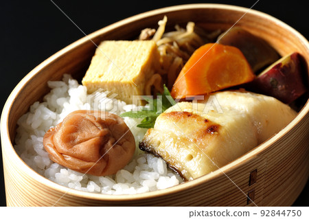 A silver cod saikyo-yaki bento with a large umeboshi on top of rice and tamagoyaki and simmered dishes, photographed against a black background. 92844750