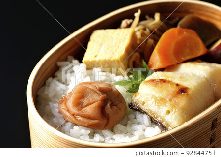 A silver cod saikyo-yaki bento with a large umeboshi on top of rice and tamagoyaki and simmered dishes, photographed against a black background. A silver cod saikyo-yaki bento with a large umeboshi on top of rice and tamagoyaki and simmered dishes, photographed against a black background. 92844751