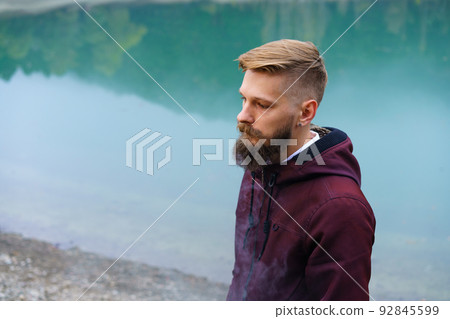 Young bearded man stood alone by lake and looked aside thoughtfully smoking Young bearded man stood alone by lake and looked aside thoughtfully smoking 92845599