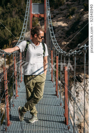 A man with a backpack on a suspension bridge against the backdrop of a mountain landscape with trees. Hiking trail pass through the hanging bridge. Travel and exploration concept 92848940