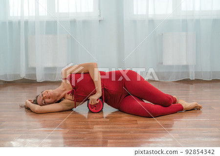 A young woman works out the fascia of the muscles with a massage roller. On the wooden floor against the background of the window in the gym 92854342