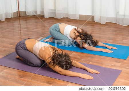 Two attractive athletic girls train asana yoga pose on mats on a light background. A group of young women stretch out in the gym. Healthy lifestyle concept Two attractive athletic girls train asana yoga pose on mats on a light background. A group of young women stretch out in the gym. Healthy lifestyle concept 92854802
