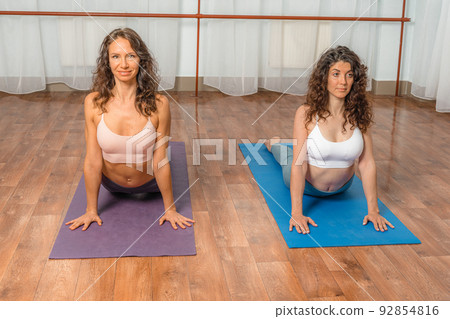 Two attractive athletic girls train asana yoga pose on mats on a light background. A group of young women stretch out in the gym. Healthy lifestyle concept 92854816