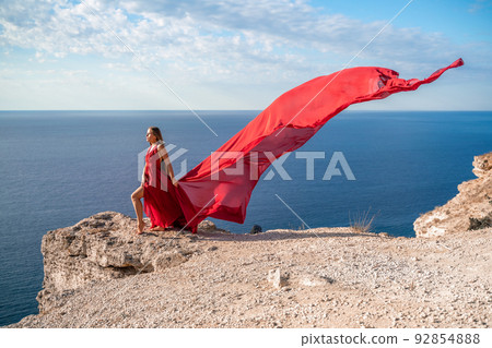 A girl with loose hair in a red dress stands on a rock rock above the sea. In the background, the sea and the rocks. The concept of travel 92854888